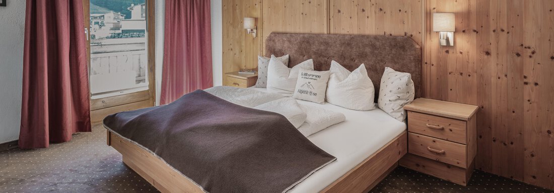 Ein gemütliches Schlafzimmer mit einem großen Bett und Holzverkleidung. Rote Vorhänge und zwei Nachttische schaffen eine einladende Atmosphäre. - Appartements Alpenrose - Huben
