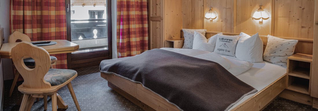Ein gemütliches Zimmer mit Holzvertäfelung, einem großen Bett und einem Fenster mit Blick auf einen Balkon. Der Raum ist schlicht eingerichtet und strahlt eine warme Atmosphäre aus. - Appartements Alpenrose - Huben