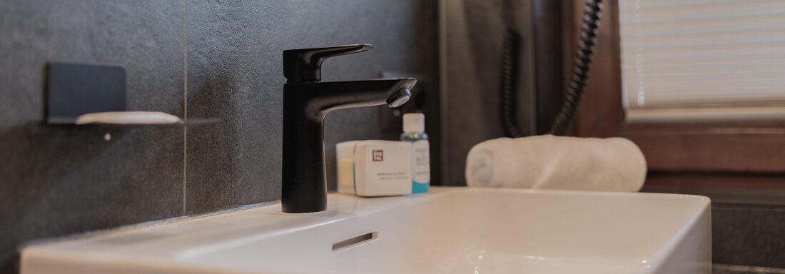 Ein modernes Waschbecken mit einem eleganten schwarzen Wasserhahn. Im Hintergrund sind Toilettenartikel und ein Handtuch sichtbar. - Appartements Alpenrose - Huben