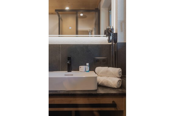 Ein modernes Badezimmer mit einem weißen Waschbecken und zwei sauberen Handtüchern. Auf dem Waschbecken sind Pflegeprodukte und eine Duschbrause zu sehen. - Appartements Alpenrose - Huben
