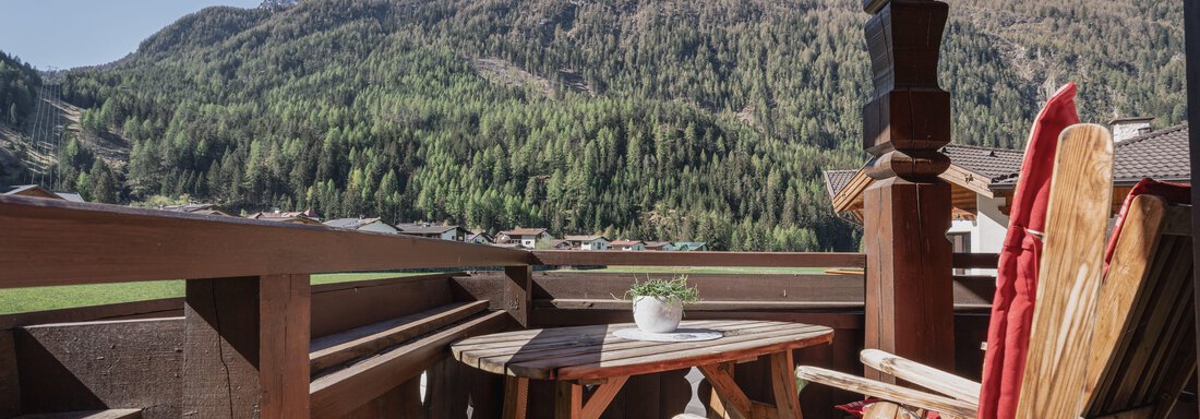 Ein gemütlicher Balkon mit einem Holztisch und Stühlen. Im Hintergrund sind beeindruckende Berge und ein grüner Wald sichtbar. - Appartements Alpenrose - Huben