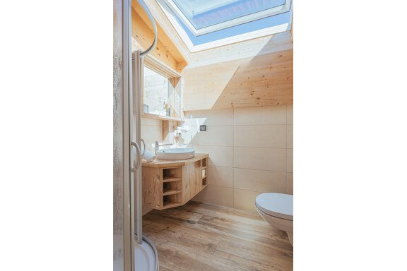 Ein modernes Badezimmer mit Holzdecken und einem großen Dachfenster. Es verfügt über ein Waschbecken, eine Dusche und ein WC in einem schlichten Design. - Appartements Alpenrose - Huben
