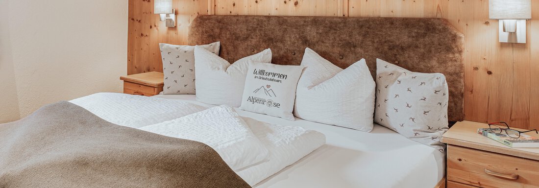 Ein gemütliches Schlafzimmer mit einem großen Bett und vielen Kissen. Die Holzwände und die sanfte Beleuchtung schaffen eine warme Atmosphäre. - Appartements Alpenrose - Huben
