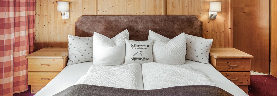 Ein gemütliches Schlafzimmer mit einem großen Bett und vielen Kissen. Die Wände sind aus hellem Holz, und es gibt zwei Nachttische neben dem Bett. - Appartements Alpenrose - Huben