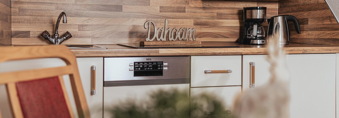 Eine moderne Küche mit Holzregalen und einer gemütlichen Atmosphäre. Auf der Arbeitsfläche steht das Wort "Dahoam" und es sind alltägliche Küchengeräte sichtbar. - Appartements Alpenrose - Huben