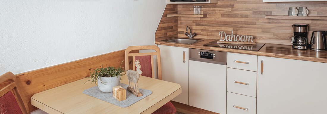 Eine moderne Küche mit Holzverkleidung und eleganten weißen Schränken. An einem Tisch stehen zwei rote Stühle und dekorative Elemente sind sichtbar. - Appartements Alpenrose - Huben