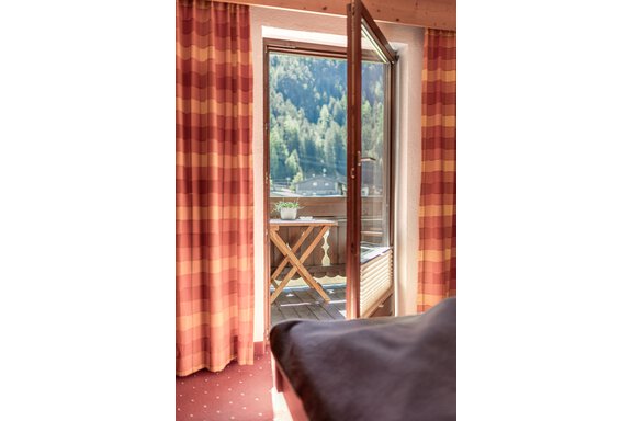 Ein gemütliches Zimmer mit offenen Fenstern, die auf einen Balkon mit Bergblick führen. Die Vorhänge sind rot gestreift und der Balkon hat einen kleinen Tisch. - Appartements Alpenrose - Huben