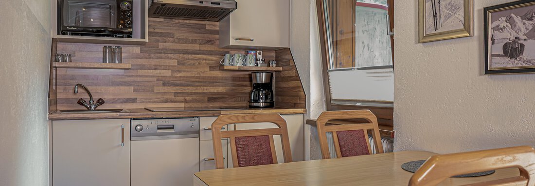 Eine moderne Küche mit Holzregalen und einem Esstisch. Es gibt eine Mikrowelle, Kaffeemaschine und Fenster für viel Tageslicht. - Appartements Alpenrose - Huben