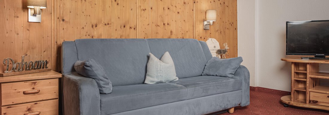 Ein gemütliches Wohnzimmer mit einem blauen Sofa und Holzverkleidung. Neben dem Sofa steht ein Holztisch mit einer Lampe und einem Fernseher. - Appartements Alpenrose - Huben