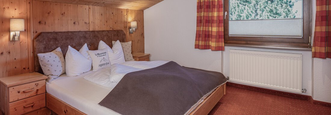 Ein gemütliches Schlafzimmer mit Holzdecke und einem großen Bett. Die Fenster bieten einen Blick auf die Natur, und die Einrichtung ist rustikal und einladend. - Appartements Alpenrose - Huben