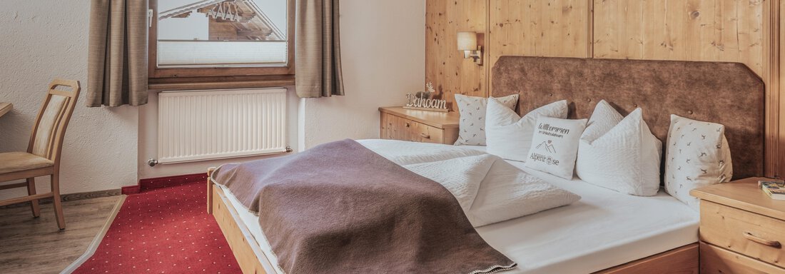 Ein gemütliches Schlafzimmer mit einem großen Bett und Holzmöbeln. Die warme Einrichtung und der Blick aus dem Fenster vermitteln eine einladende Atmosphäre. - Appartements Alpenrose - Huben