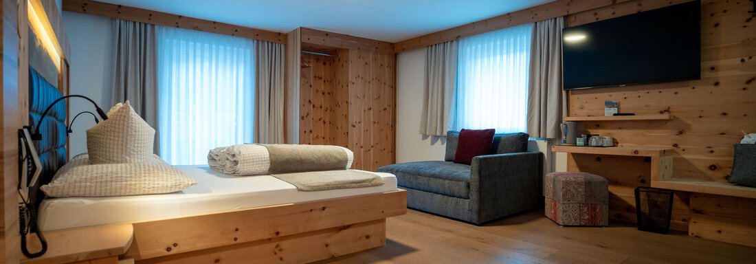 Alpenblick neu-068 A modern room with wood paneling and windows that let in daylight. There is a bed, a seating area, and a television. - Genusshotel Alpenblick - Huben