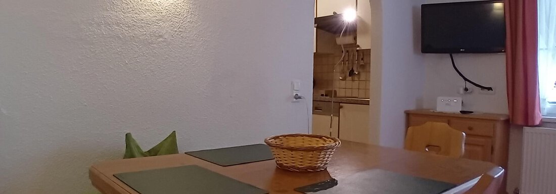 A cozy dining area with a wooden regular table and green seats. The room has warm lighting and a television on the wall. - Haus Christoph im Herzen des Ötztals - Huben