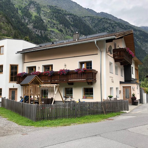 Ein modernes, mehrstöckiges Haus in bergiger Umgebung. Es hat einen Balkon mit Blumen und eine kleine Spielplatzanlage vor dem Haus. - Apartment Felix - Huben