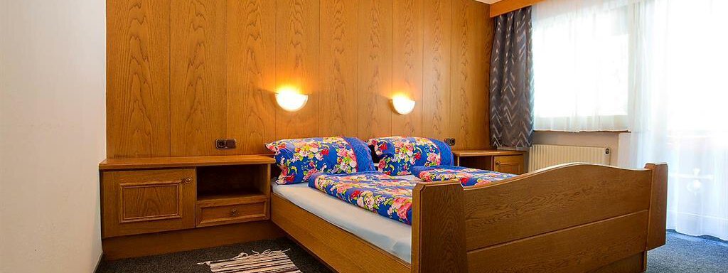 Ein einfaches Schlafzimmer mit einem hölzernen Bett und bunten Bettwäsche. Die Wände sind hellbraun, und es gibt einen kleinen Teppich auf dem Boden. - Haus Max - Huben