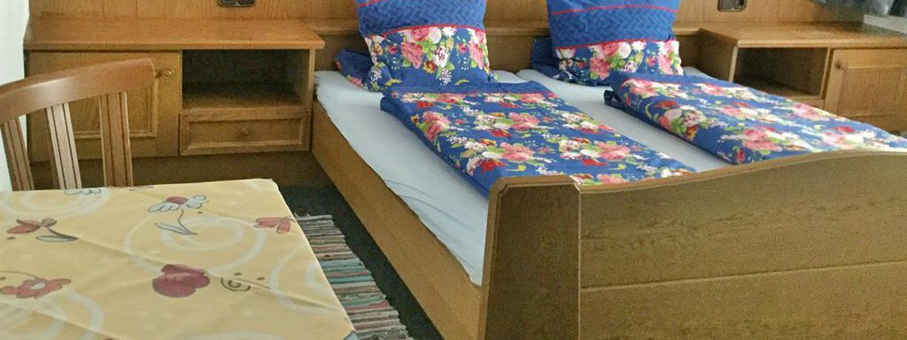 Ein gemütliches Zimmer mit zwei Einzelbetten und bunten Bettdecken. Am Boden steht ein Tisch mit einer blümchenmusterhaften Tischdecke. - Haus Max - Huben