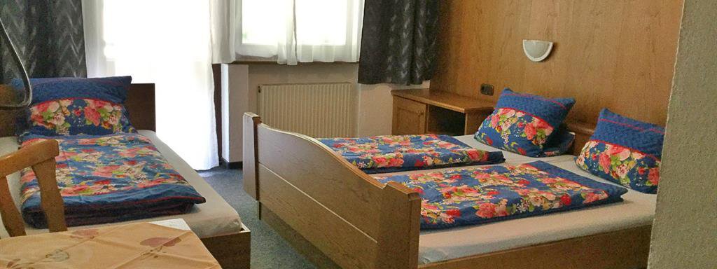 Ein einfaches Zimmer mit zwei Einzelbetten und einem Doppelbett. Bunte Bettwäsche und große Fenster sorgen für eine helle Atmosphäre. - Haus Max - Huben