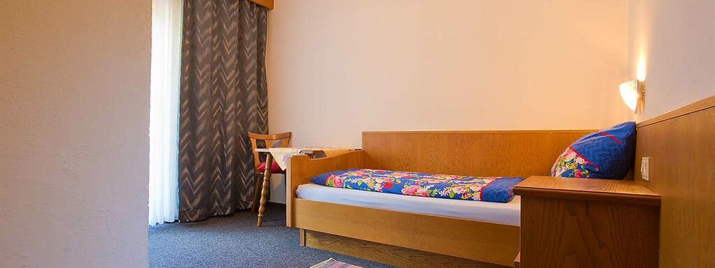 Ein einfaches Zimmer mit einem Einzelbett und einem kleinen Tisch. Es gibt Vorhänge und einen Teppich auf dem Boden. - Haus Max - Huben
