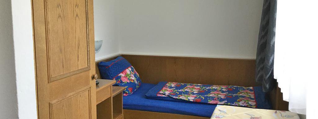 Ein einfaches Zimmer mit einem Einzelbett und bunten Kissen. Ein Tisch und ein Stuhl stehen neben dem Bett, und es gibt ein Fenster mit Vorhängen. - Haus Max - Huben