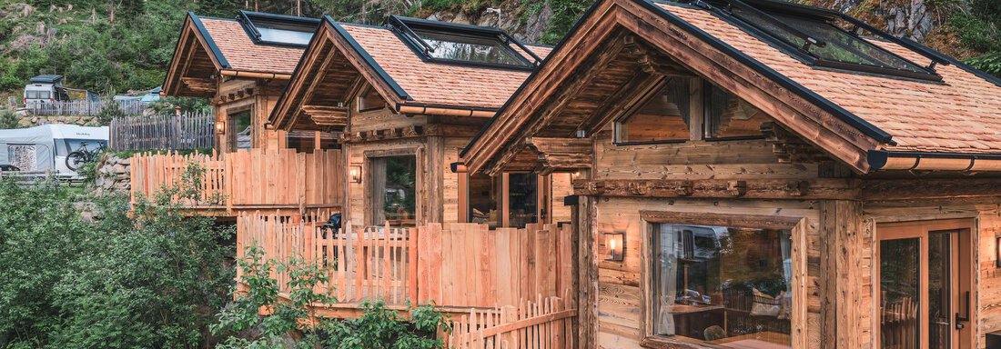Drei gemütliche Holzhütten stehen umgeben von Bäumen. Die Häuser haben große Fenster und eine einladende Veranda. - Naturcamping Kuprian - Ötztal - Huben
