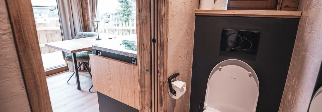 Ein modernes Bad mit WC und einem hellen, rustikalen Ambiente. Im Hintergrund ist ein kleiner Arbeitsbereich mit einem Tisch und einem Fenster zu sehen. - Naturcamping Kuprian - Ötztal - Huben