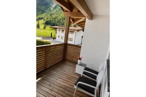 Ein gemütlicher Balkon mit Holzgeländer und zwei weißen Stühlen. Im Hintergrund sind grüne Berge und ein weißes Haus zu sehen. - ONO Apartments - Huben