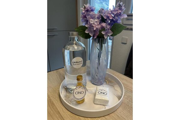 Ein Dekotablett mit einer Glasflasche Wasser, einer Vasen mit lila Blumen und einem kleinen Fläschchen mit dem Aufdruck „ONO“. Dazu liegt eine Packung im gleichen Design. - ONO Apartments - Huben