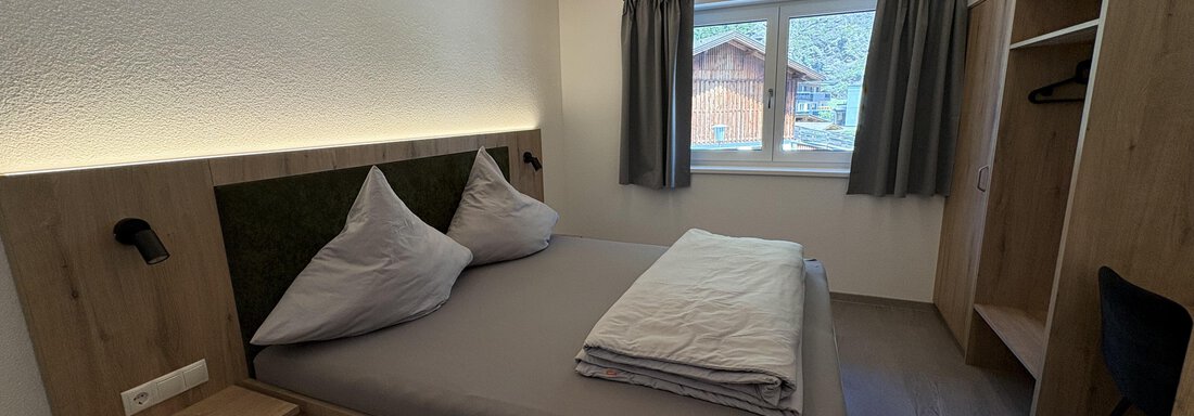 A cozy bedroom with wooden decor and a large bed. The windows let in plenty of natural light and offer a view outside. - 6444 Apartment - Längenfeld