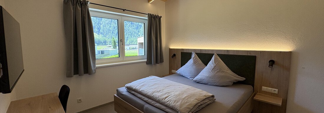 A cozy bedroom with a large bed and wooden decor. A lot of daylight is visible through the window. - 6444 Apartment - Längenfeld