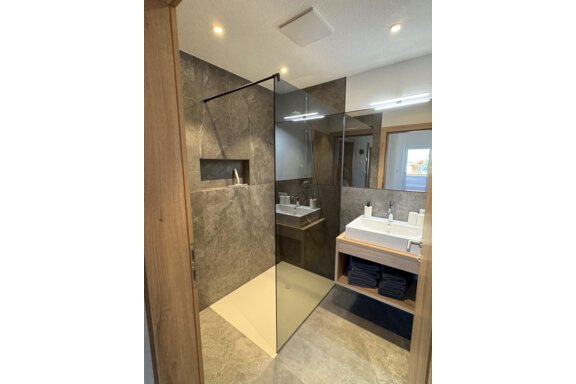 A modern bathroom with an elegant glass shower and a large sink area. The walls are decorated with gray tiles, and there is a relaxed atmosphere. - 6444 Apartment - Längenfeld
