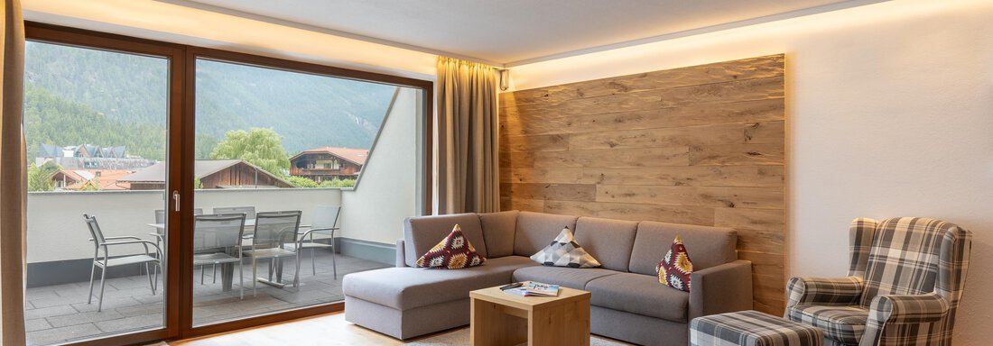 Ein modernes Wohnzimmer mit grauem Sofa und gemütlichem Sessel. Große Fenster bieten einen Blick auf die Berge und einen Balkon. - Alpenappartement Central Valley Penthouse - Längenfeld