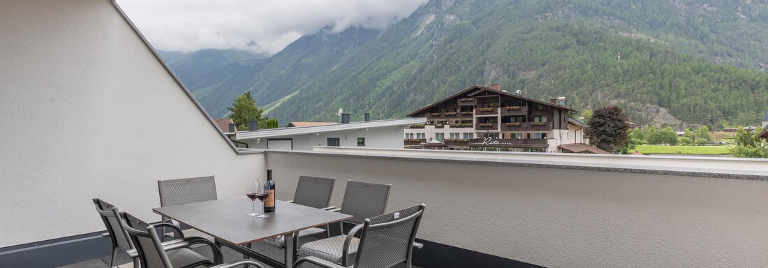 Ein Balkon mit einem Tisch und Stühlen, umgeben von Bergen und Wolken. Im Hintergrund ist ein Gebäude zu sehen. - Alpenappartement Central Valley Penthouse - Längenfeld