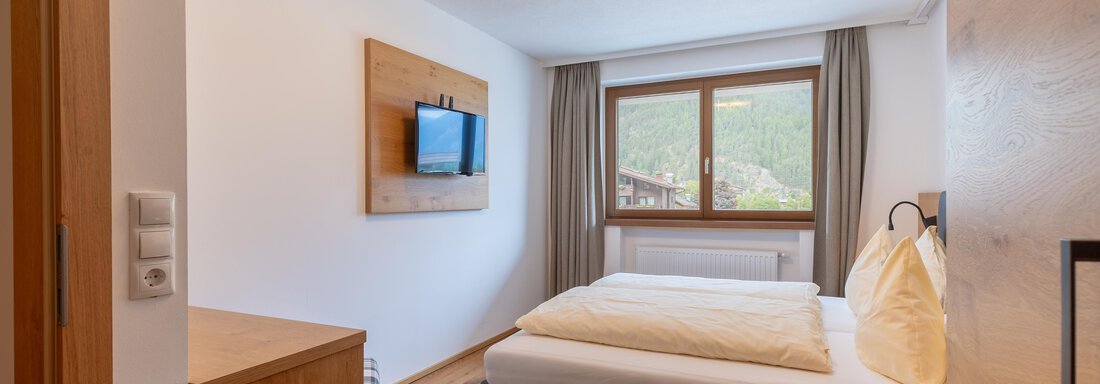 Ein gemütliches Schlafzimmer mit einem Doppelbett und einem Fernseher an der Wand. Der Raum hat große Fenster mit Blick auf die Berge und helles Holzdekor. - Alpenappartement Central Valley Penthouse - Längenfeld