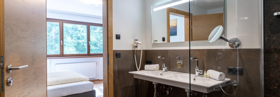 Ein modernes Badezimmer mit einer Glasdusche und einem Doppelwaschbecken. Im Hintergrund ist ein Schlafzimmer mit einem großen Bett und Fenstern zu sehen. - Alpenappartement Central Valley Penthouse - Längenfeld
