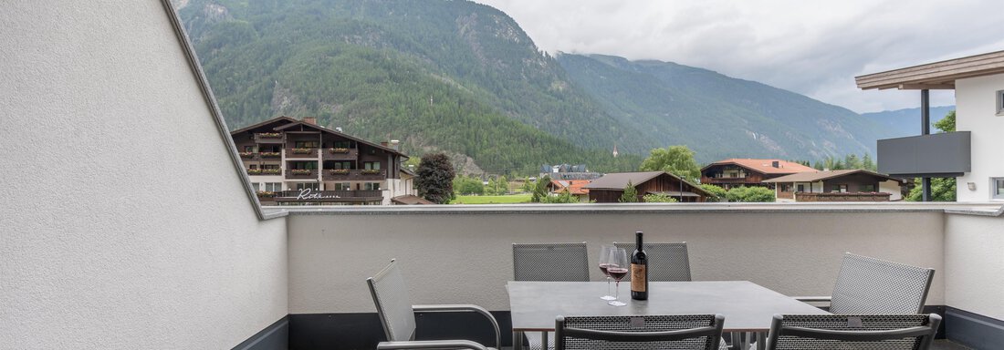 Eine Terrasse mit einem Tisch und Stühlen, auf dem eine Flasche Wein steht. Im Hintergrund sind Berge und Wolken zu sehen. - Alpenappartement Central Valley Penthouse - Längenfeld