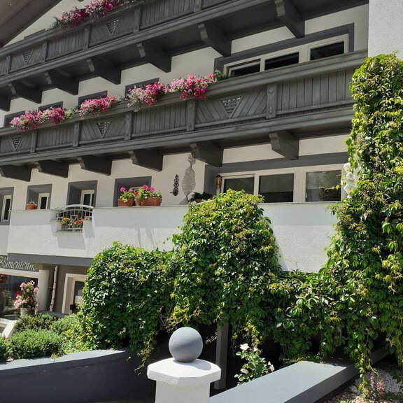 A charming building with balconies, adorned with colorful flowers. Lush greenery surrounds the entrance, creating an inviting atmosphere. - Apart Alpenland Maurer - Längenfeld