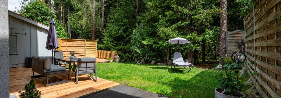 A quiet garden with green lawn and tall trees. There is a wooden terrace with seating options and a deck chair. - Apart Conny - Längenfeld