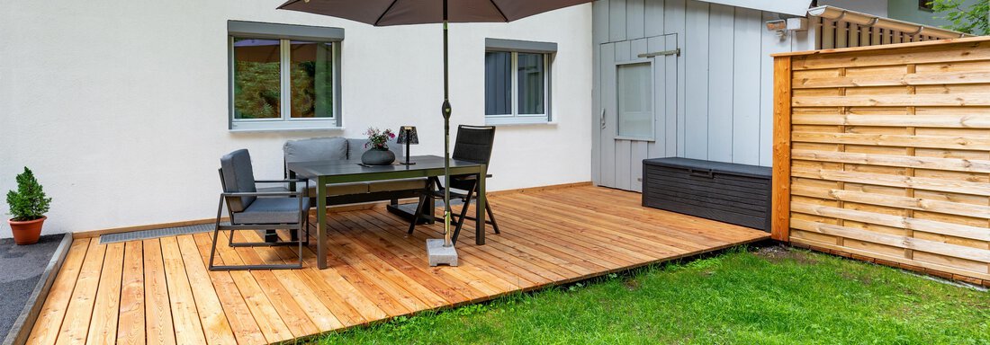 A cozy terrace with a wooden floor and a table under a sun umbrella. In the background, a wall and a small garden can be seen. - Apart Conny - Längenfeld