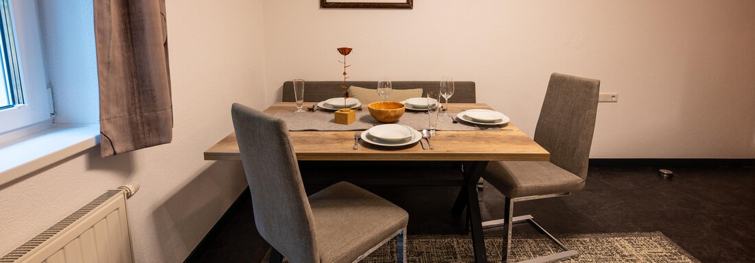 A cozy dining area with a wooden table and stylish chairs. The table is set with dishes and a decorative fruit bowl. - Apart Conny - Längenfeld