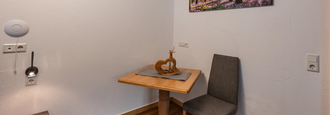 A small dining area with a wooden table and a chair. On the wall hangs a picture of a forest with sunlight. - Apart Conny - Längenfeld