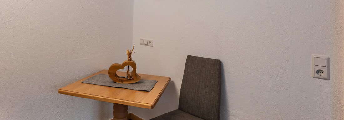 A small dining area with a wooden chair and a table. On the wall hangs a picture of a forest with sun rays. - Apart Conny - Längenfeld