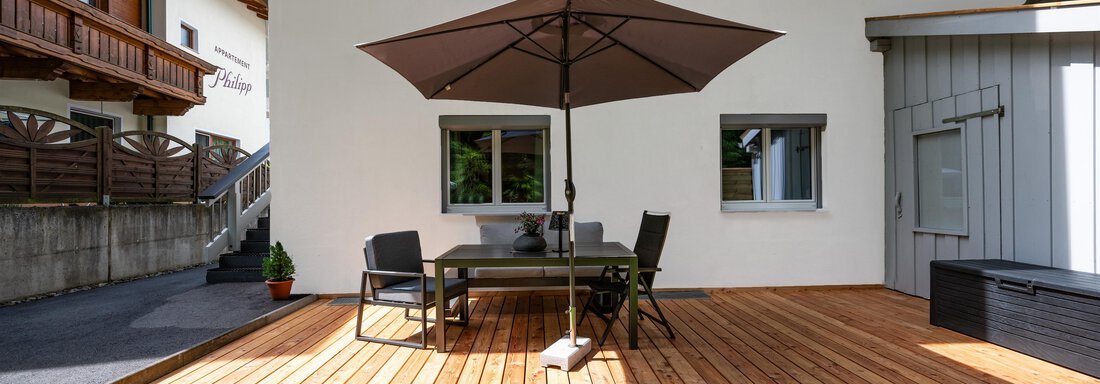A wooden terrace with a table and chairs, surrounded by green grass. A sunshade provides shade, and buildings are visible in the background. - Apart Conny - Längenfeld