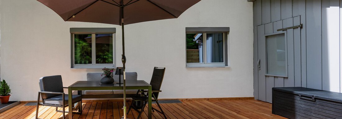 A terrace with a large sun umbrella, a table, and chairs. In the background, there is a white house and the lawn is green. - Apart Conny - Längenfeld