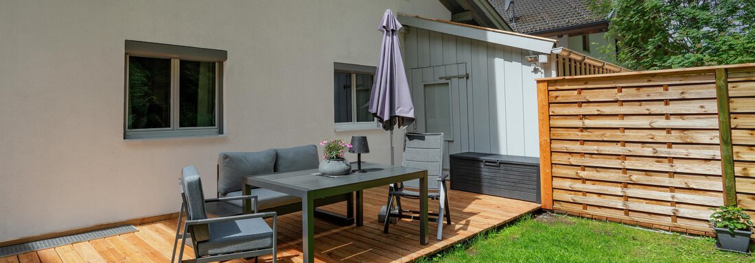 A cozy terrace with a table and chairs. In the background, there is a wooden fence and green grass. - Apart Conny - Längenfeld