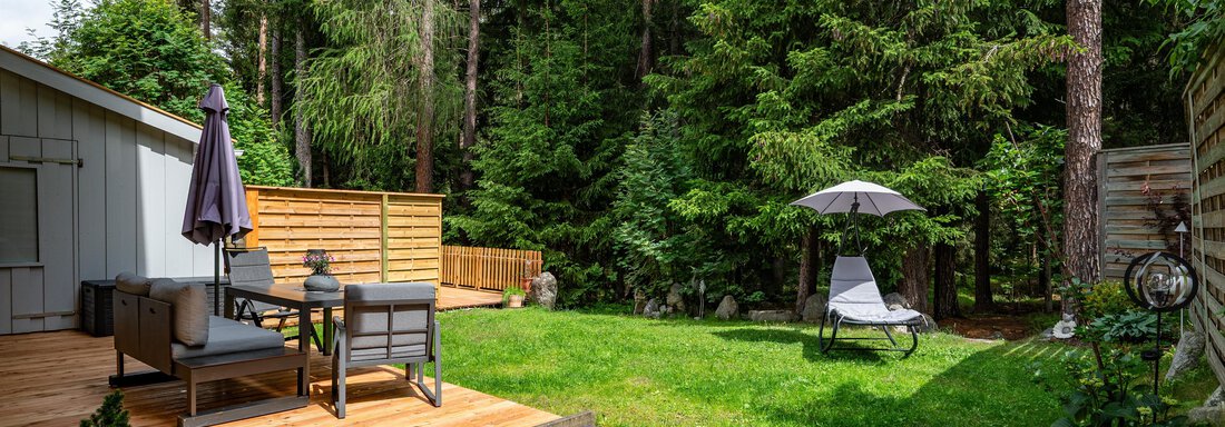 A quiet garden with a wooden terrace and a comfortable lounge chair. Surrounded by tall trees, it offers a relaxed atmosphere. - Apart Conny - Längenfeld