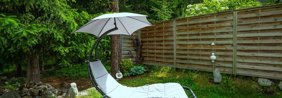 A garden lounge chair with a sun umbrella stands on green grass. Surrounded by trees and a wooden fence, it offers a quiet retreat. - Apart Conny - Längenfeld