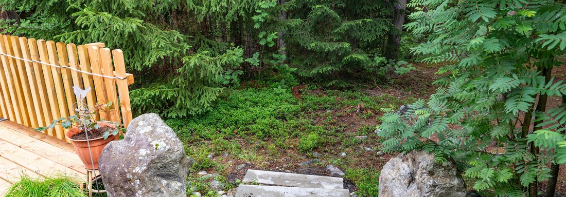 A small garden path made of stones leads into a green, wooded area. In the foreground, there is a wooden terrace with plants and stones. - Apart Conny - Längenfeld