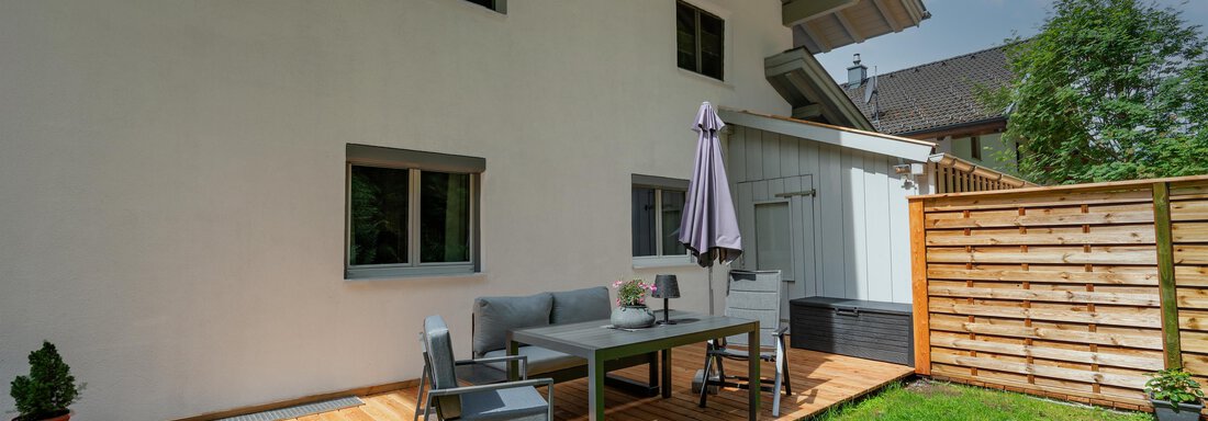 A cozy terrace with a wooden floor and a table as well as chairs. In the background, there is a modern house and a privacy fence. - Apart Conny - Längenfeld