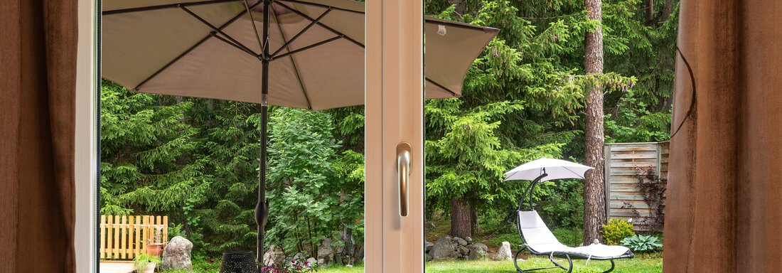 A view through a window onto a garden with a sun umbrella and loungers. In the foreground, there is a heart-shaped wooden decoration. - Apart Conny - Längenfeld