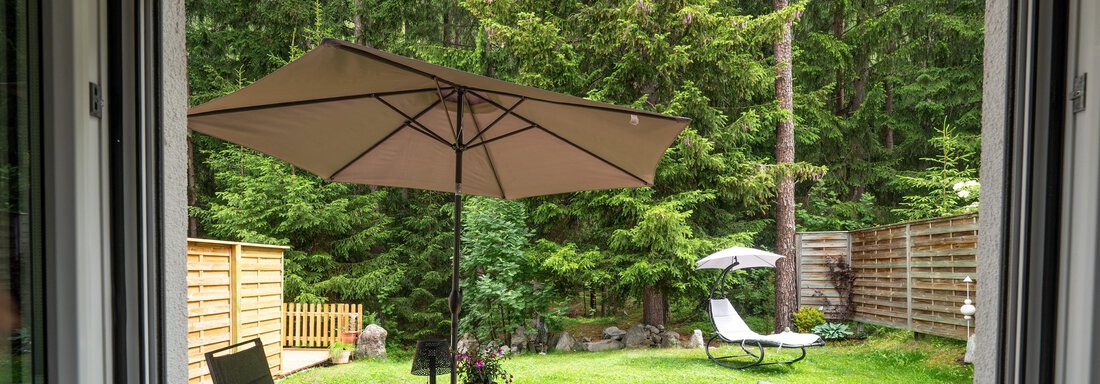 A beautiful garden with a table, chairs, and a sun umbrella. In the background, there are trees and a lounge chair. - Apart Conny - Längenfeld